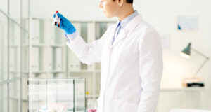 Young female researcher or chemist in whitecoat and gloves holding two flasks with samples of blood over glass cage with white mice